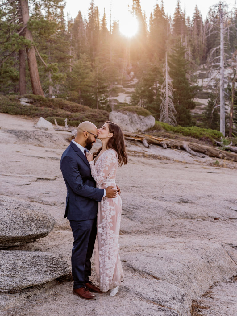 Bride kisses groom on the forehead with the sun setting over the trees in Yosemite
